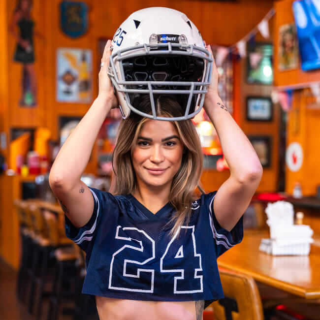 Woman in a cropped football jersey lifting a helmet inside a sports bar.