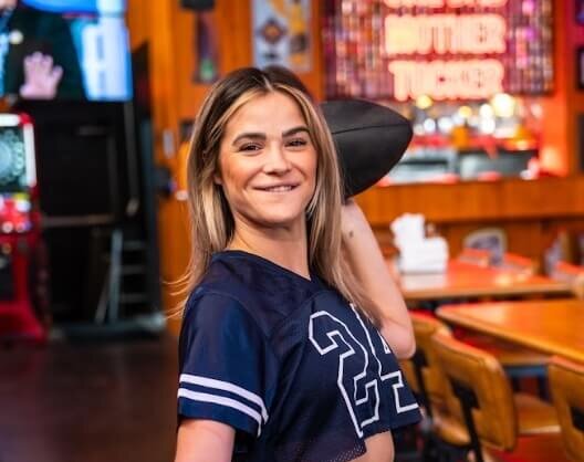 Woman wearing a navy cropped football jersey holds a football and smiles inside a rustic sports bar with high-top chairs and neon signage reading “Mother Tucker.”