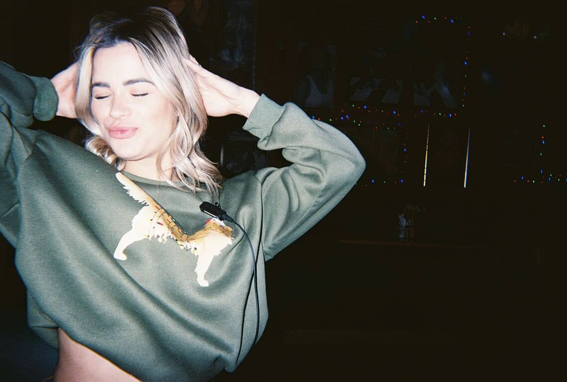 Young woman in a cropped green sweatshirt posing playfully with eyes closed at a cozy bar with moody lighting and string lights in the background candid nightlife photography.