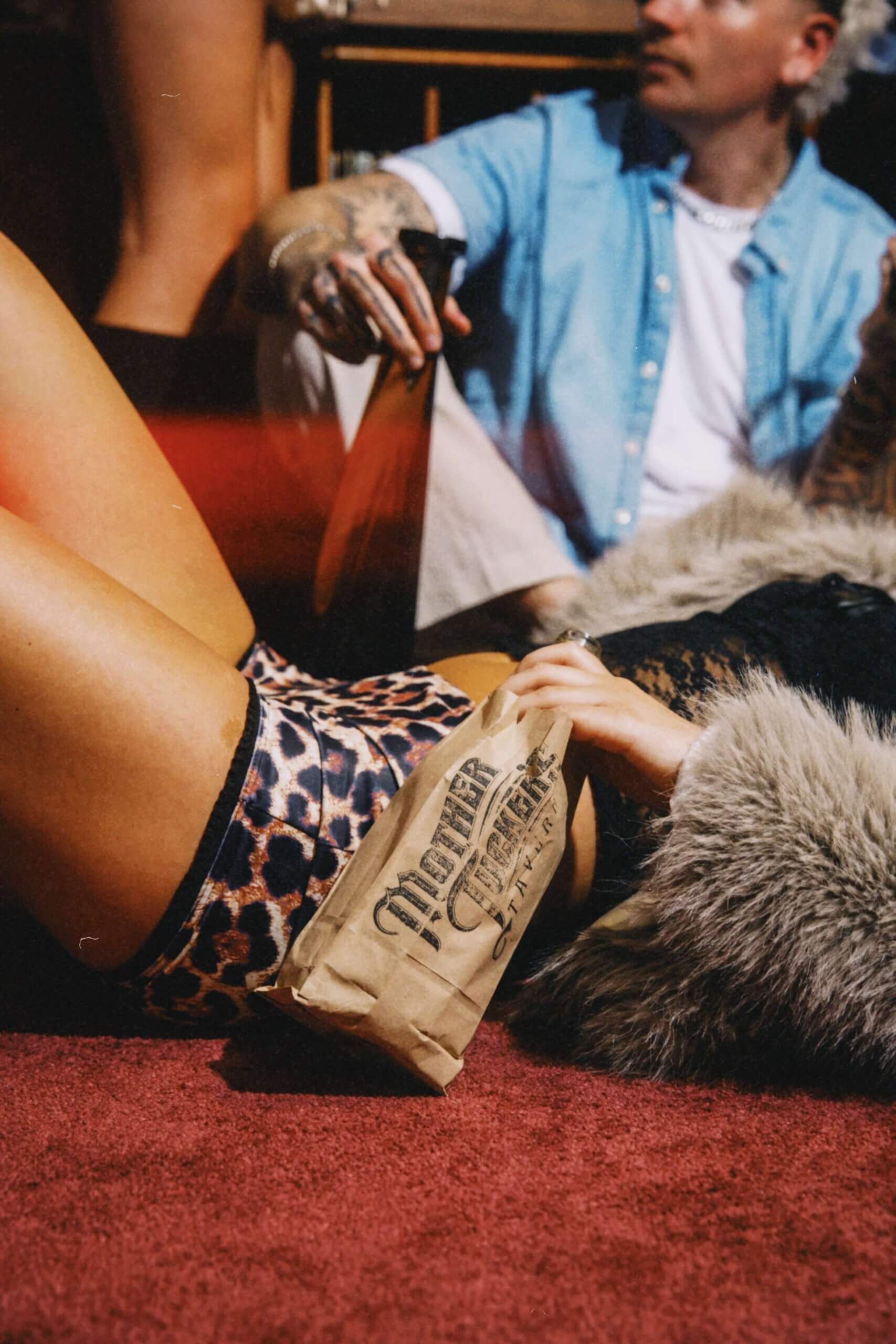 Close-up of a person lounging on a red carpet, wearing leopard-print shorts and a fur coat, holding a brown paper bag with the “Mother Tuckers Tavern” logo.