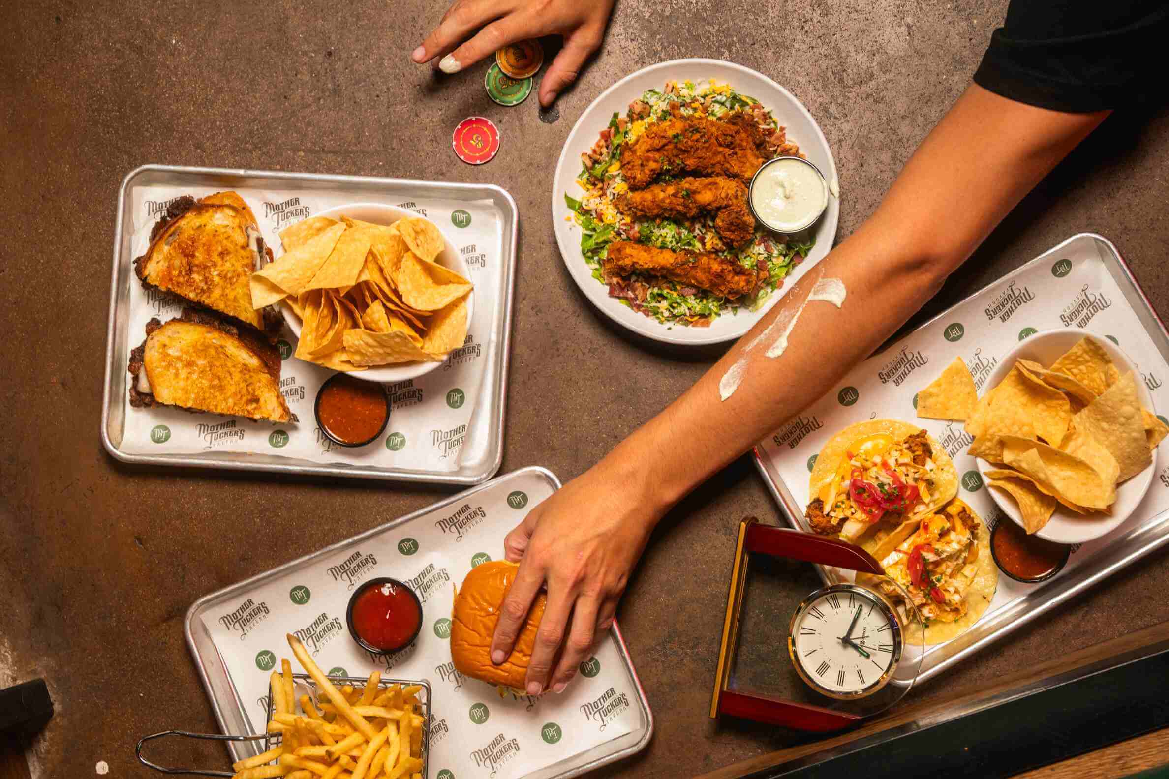 A vibrant top-down view of a delicious spread at Mother Tucker's featuring tacos, grilled sandwiches, fries, chips with salsa, a chicken tender salad, and a juicy burger perfect for casual dining or game night.
