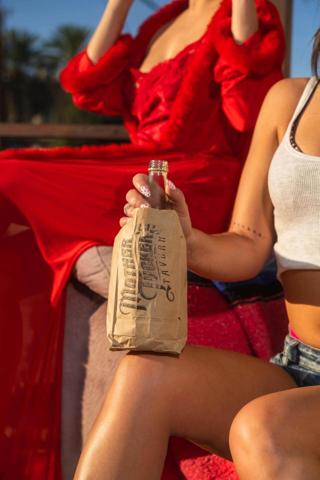 A warm, sunlit close-up of a woman holding a drink wrapped in a Mother Tucker’s Tavern paper bag. Casual summer vibes meet bold style with playful nail art and a relaxed outdoor setting.