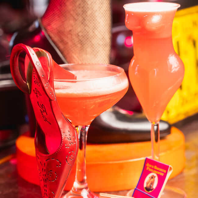 Two pink cocktails in uniquely shaped glasses, one featuring a red high heel garnish and the other in a torso-shaped glass, set against a sultry bar background with fishnet stockings and dim lighting.