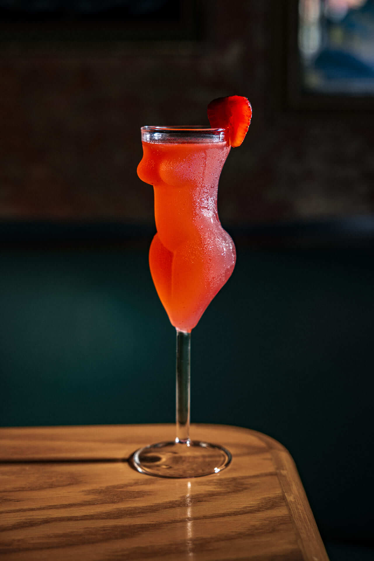 Red cocktail served in a sculpted glass shaped like a woman's body with a strawberry garnish, placed on a wooden table.