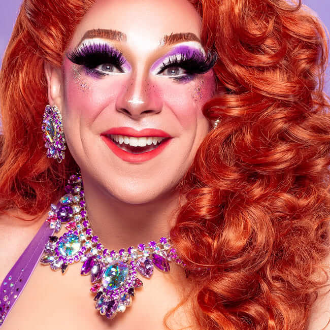Smiling drag performer with bright red hair, bold purple eye makeup, and sparkling jeweled necklace against a colorful backdrop.