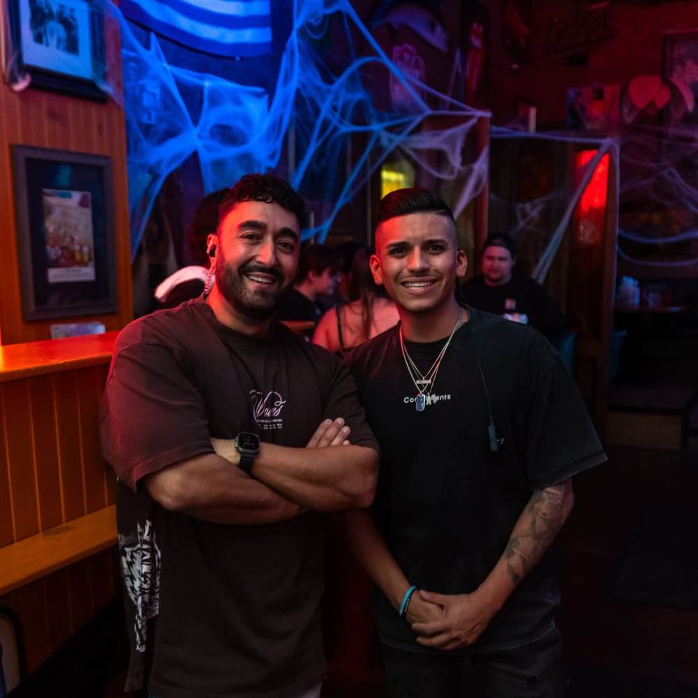 Two smiling men standing together inside a dimly lit bar decorated with blue spiderweb lights, wooden paneling, and framed pictures on the wall.
