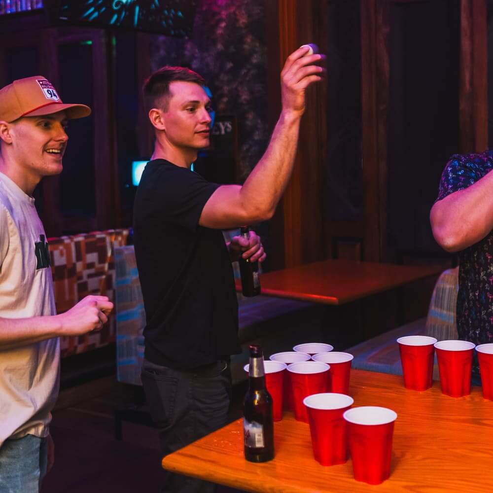 Two men playing beer pong in a dimly lit bar, with red cups arranged on a wooden table and one player tossing a ping-pong ball.