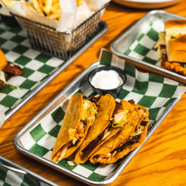 Three crispy tacos filled with shredded meat and toppings served on a metal tray lined with green-and-white checkered paper, with a small cup of white dipping sauce beside them.