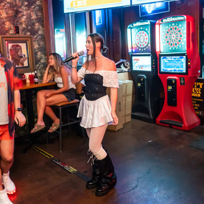 Woman singing karaoke in a bar with dart machines and seated guests in the background
